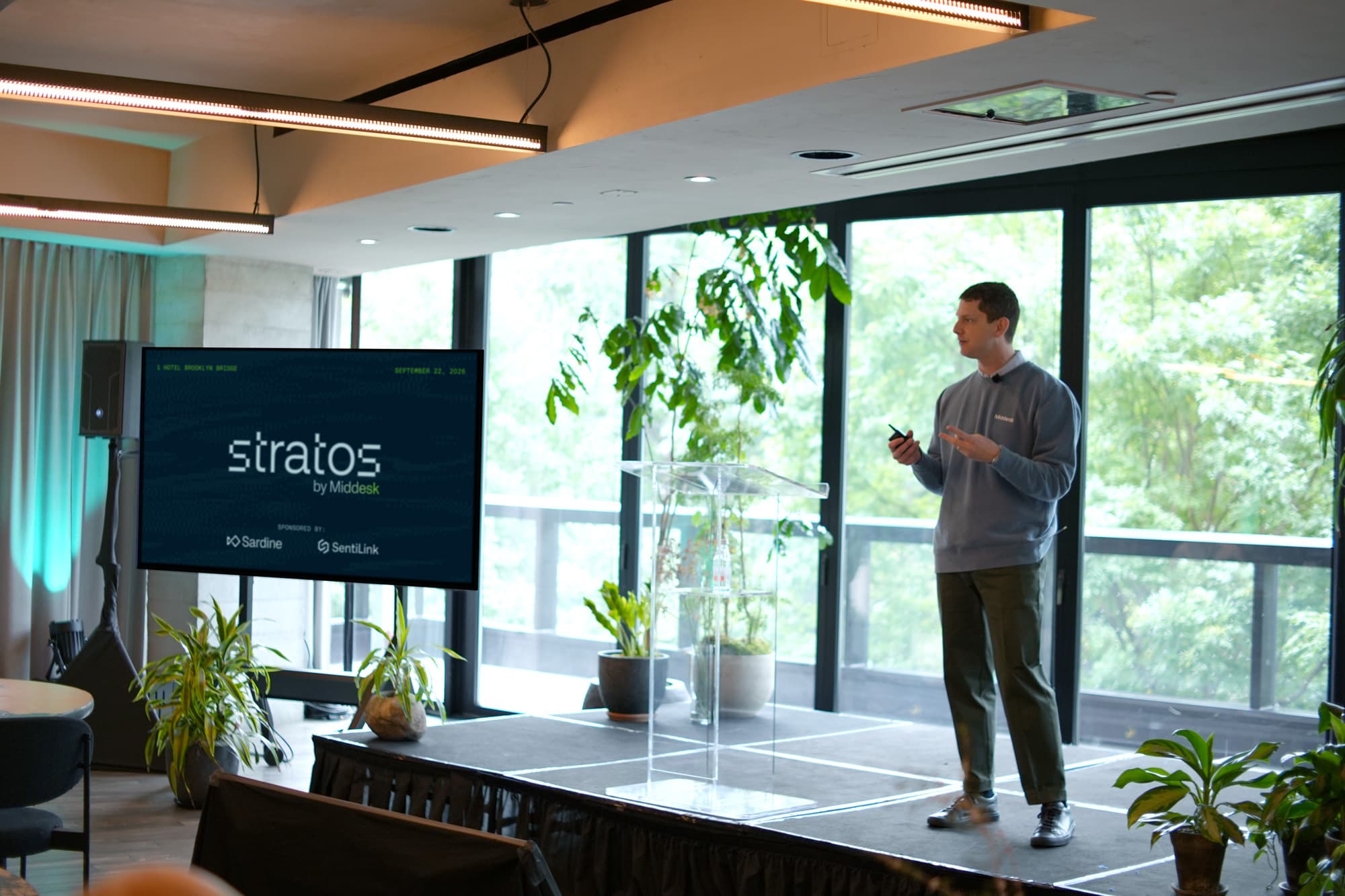 A speaker gives a presentation next to a screen displaying "Stratos by Middesk" in a well-lit room with potted plants and large windows.